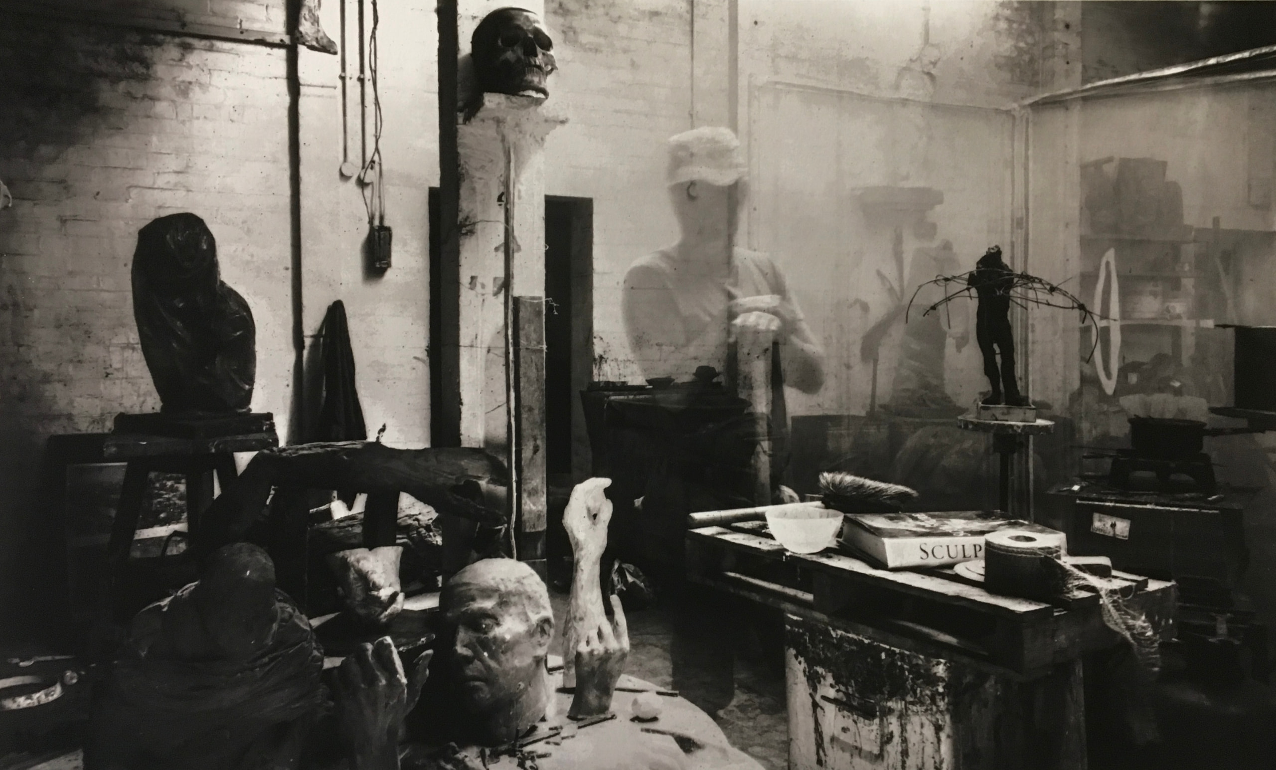Black-and-white studio interior with a translucent figure among sculpted heads, a skull and worktables
